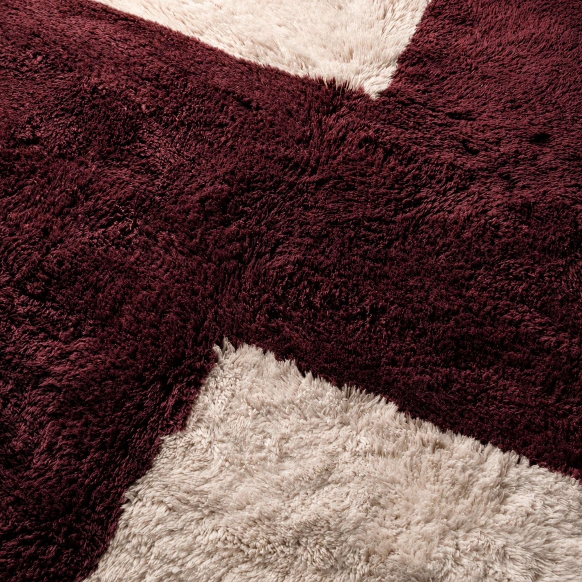 Close-up detail of handmade burgundy and cream Moroccan wool rug showing dense shag pile, soft texture, natural wool variation, and sculptural geometric intersection.