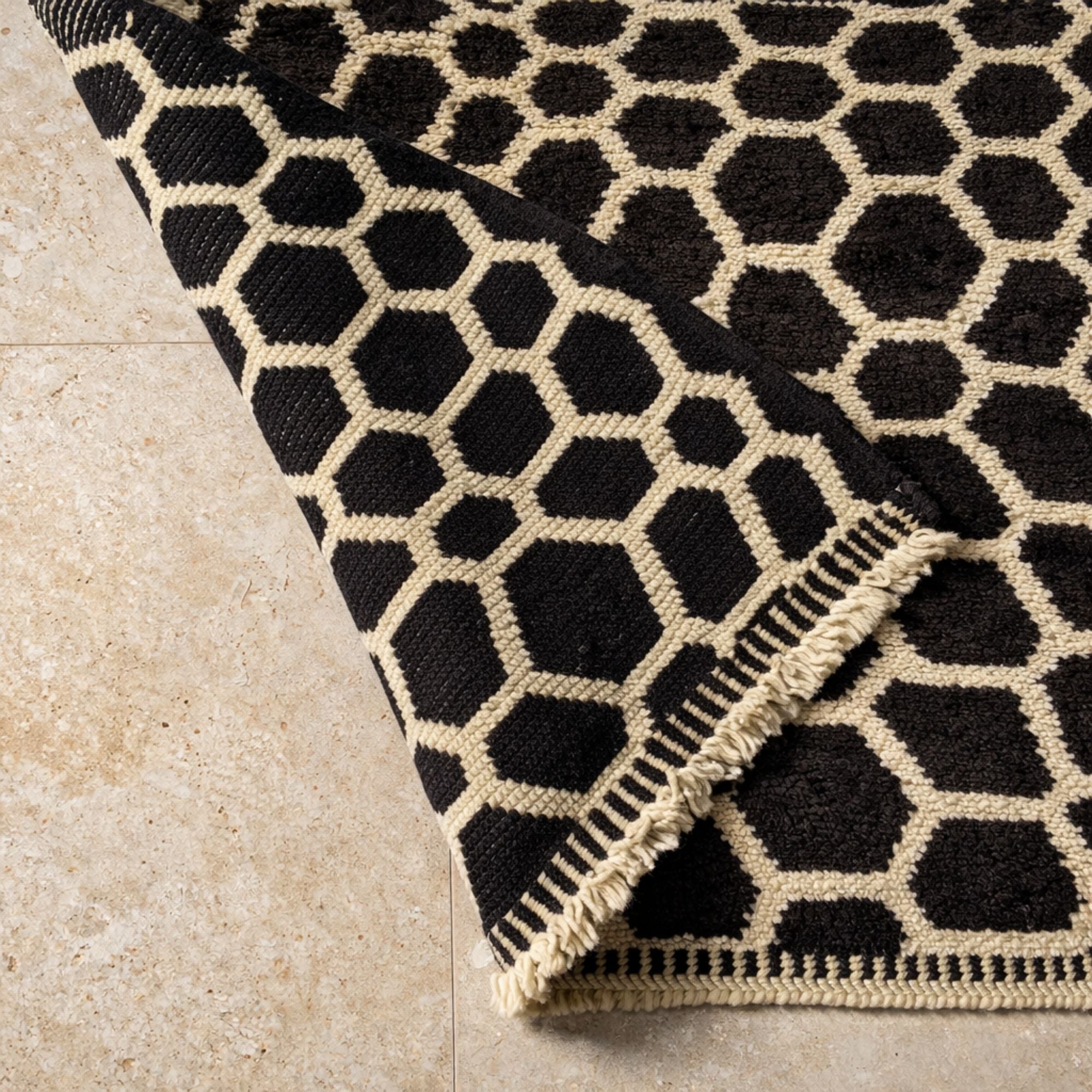 Close-up of a handcrafted Moroccan wool rug folded to reveal construction and texture. Black geometric pebble motifs contrast against an ivory base with handwoven border detailing and natural wool fringe.