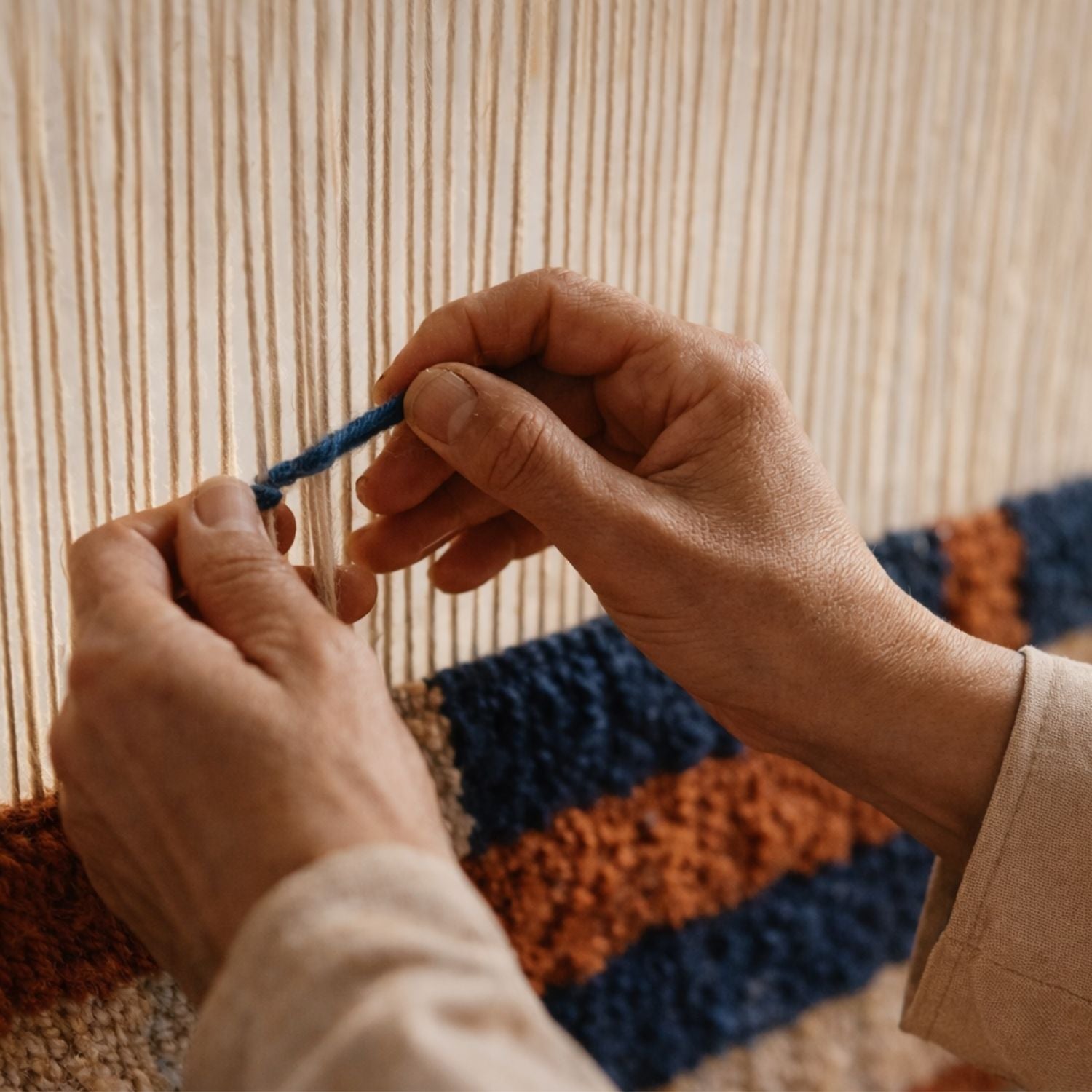 Detailed close-up of a Moroccan Berber artisan weaving a handmade wool rug on a traditional loom, tying blue yarn into off-white vertical threads with visible texture and craftsmanship